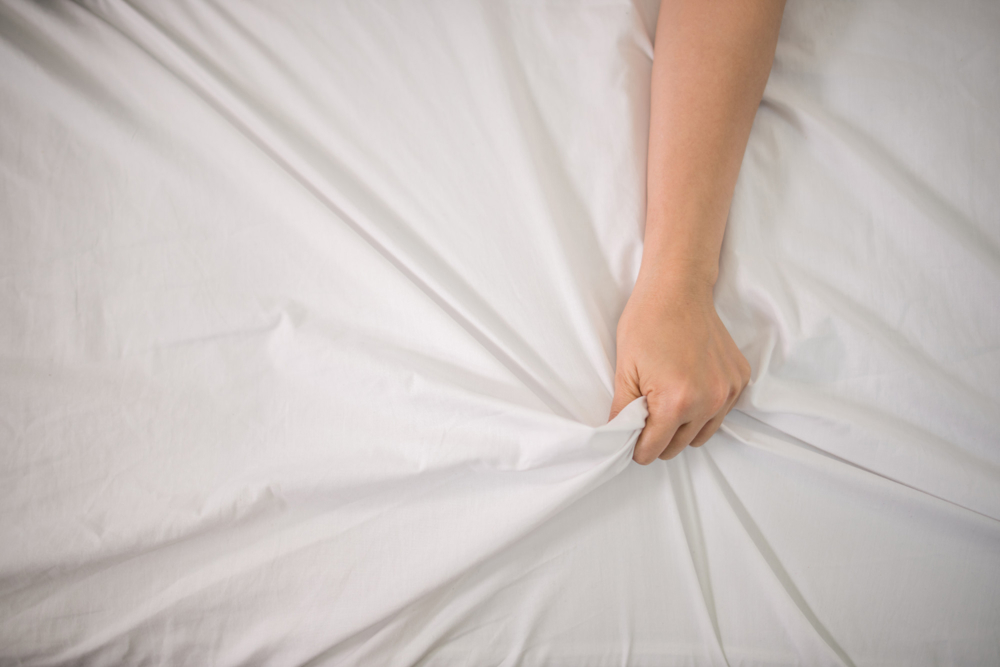 A hand grabbing the white sheets of a bed in an orgasmic way.